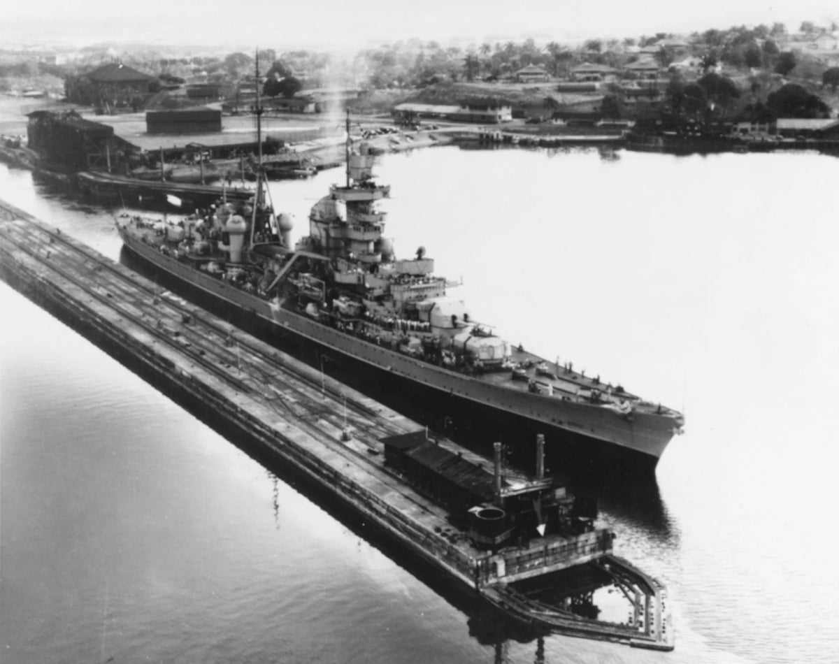 Black and white historical photograph of a large military battleship docked in a harbor, with industrial buildings and trees visible in the background.
