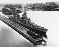 Black and white historical photograph of a large military battleship docked in a harbor, with industrial buildings and trees visible in the background.