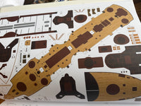 A close-up of a model kit decal sheet featuring a large wooden-texture hull panel and various smaller brown and black detail stickers on a white backing card.