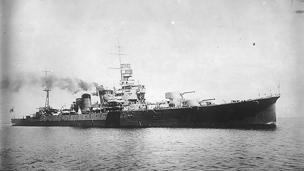 Black and white historical photograph of a large military battleship at sea, with smoke rising from its funnels and open ocean in the foreground.