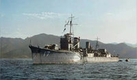 A large gray military ship sailing on calm blue water with mountains in the background under a pale sky.