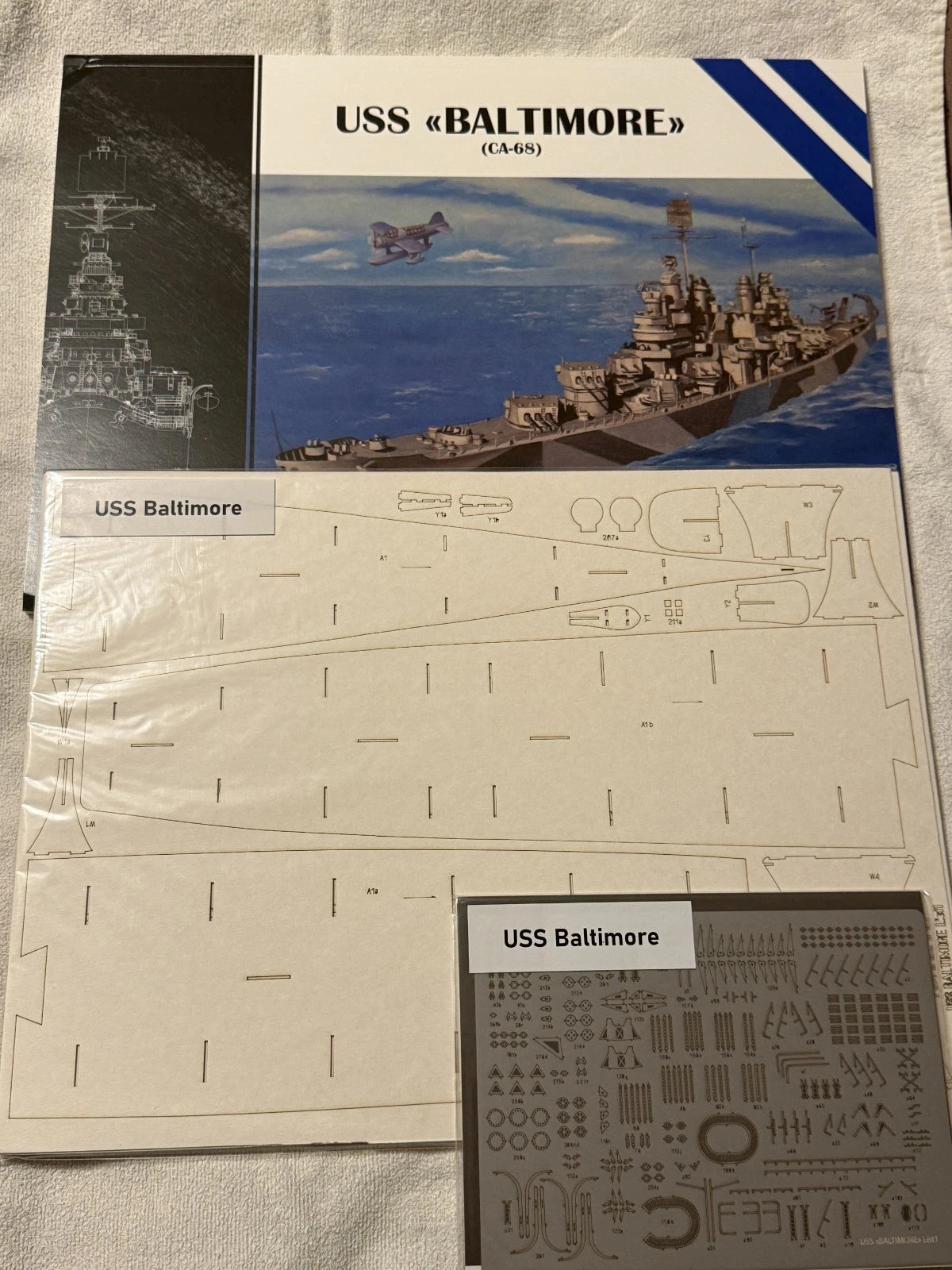 Model kit parts and packaging for a USS Baltimore (CA-65) model ship, including a printed box cover, flat laser-cut panel pieces, and a sheet of decals, laid out on a light fabric background.