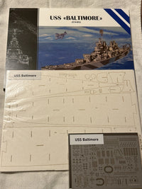 Model kit parts and packaging for a USS Baltimore (CA-65) model ship, including a printed box cover, flat laser-cut panel pieces, and a sheet of decals, laid out on a light fabric background.