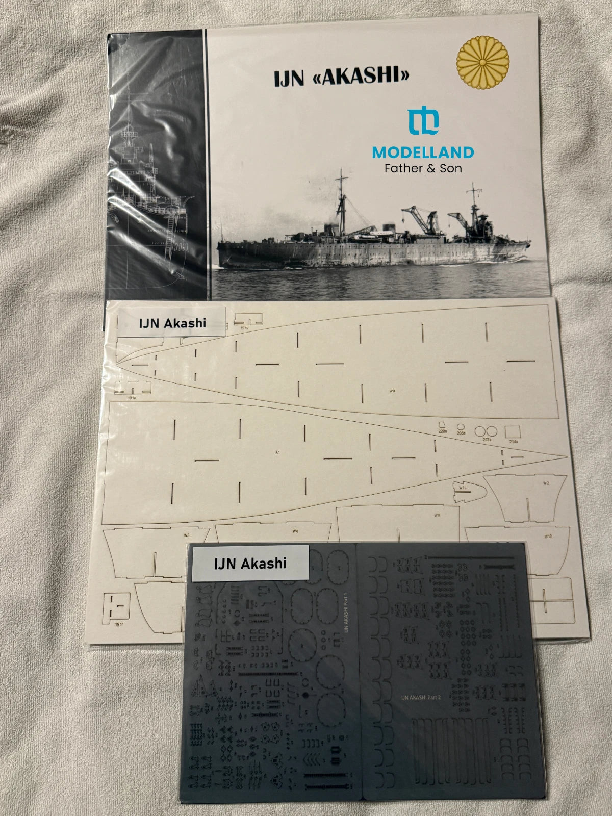 UN Akashi model kit parts and instruction materials laid out on a light fabric surface, including a printed instruction sheet, a dark gray decals sheet labeled 'UN Akashi,' and the front cover of the model kit box showing the ship and Modelland branding.