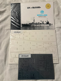 UN Akashi model kit parts and instruction materials laid out on a light fabric surface, including a printed instruction sheet, a dark gray decals sheet labeled 'UN Akashi,' and the front cover of the model kit box showing the ship and Modelland branding.