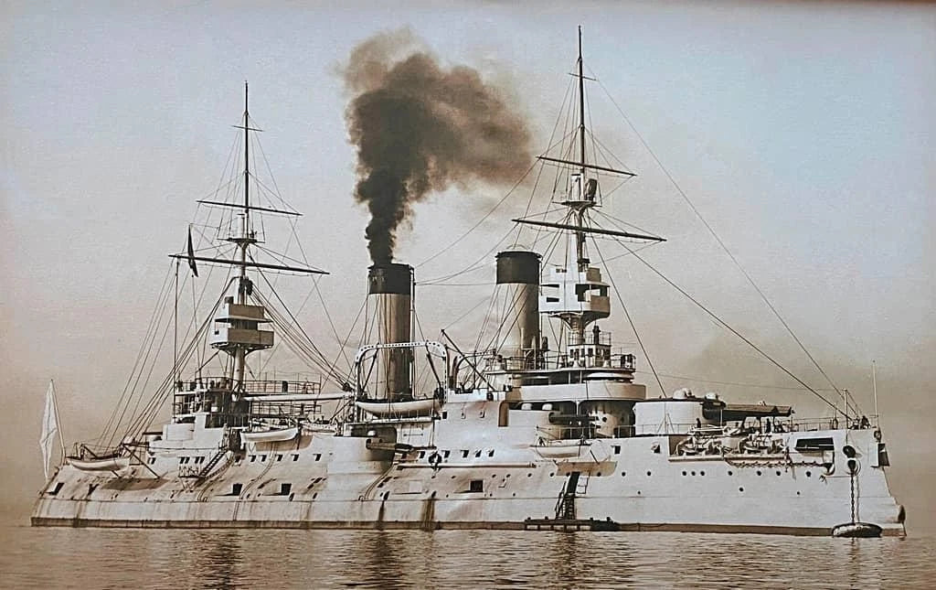 A vintage-toned photograph of a large naval battleship at sea, with smoke billowing from its funnels and a pale sky in the background.