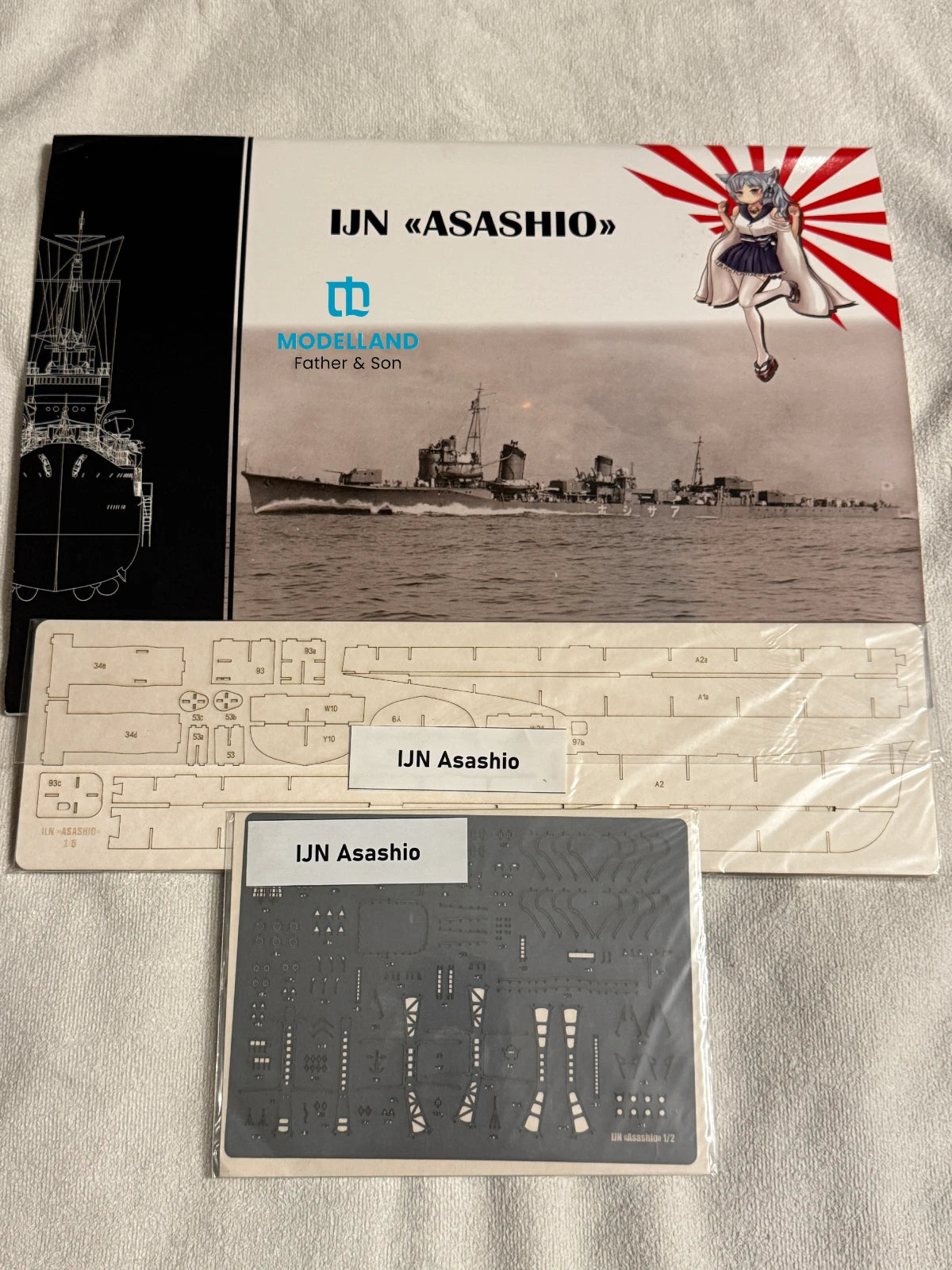 Model kit packaging and parts laid out on a light fabric surface. The set includes a printed cover card for a UJN Asashio model, a large beige plastic sheet with cutout parts, and a dark gray decal sheet under clear plastic.