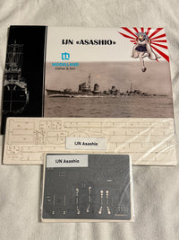 Model kit packaging and parts laid out on a light fabric surface. The set includes a printed cover card for a UJN Asashio model, a large beige plastic sheet with cutout parts, and a dark gray decal sheet under clear plastic.