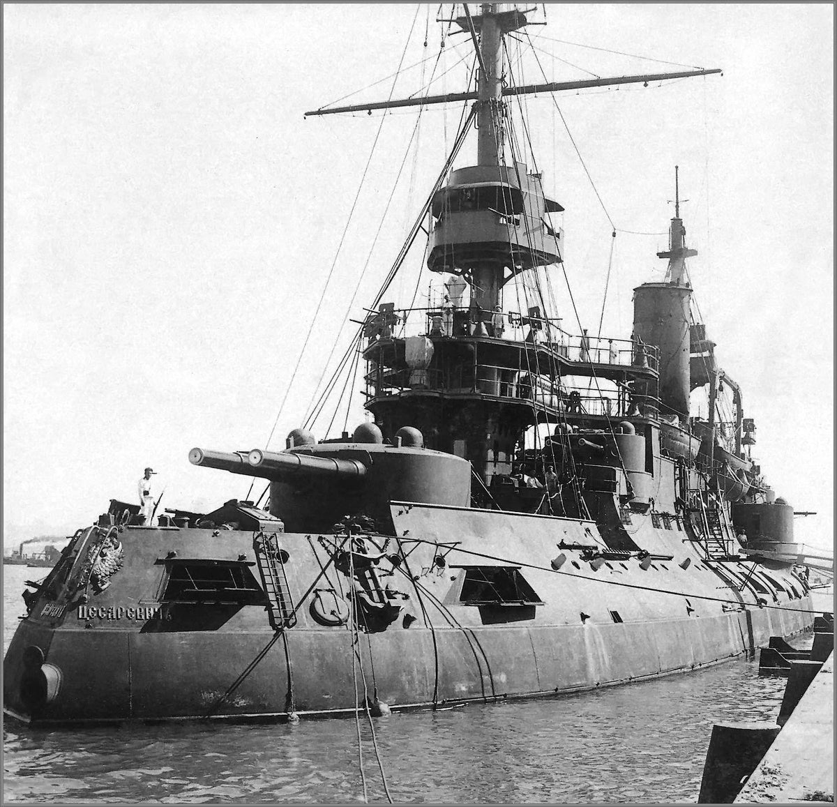 Black and white historical photograph of a large military battleship docked in water, with visible masts, gun turrets, and harbor surroundings.