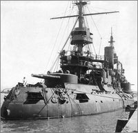 Black and white historical photograph of a large military battleship docked in water, with visible masts, gun turrets, and harbor surroundings.