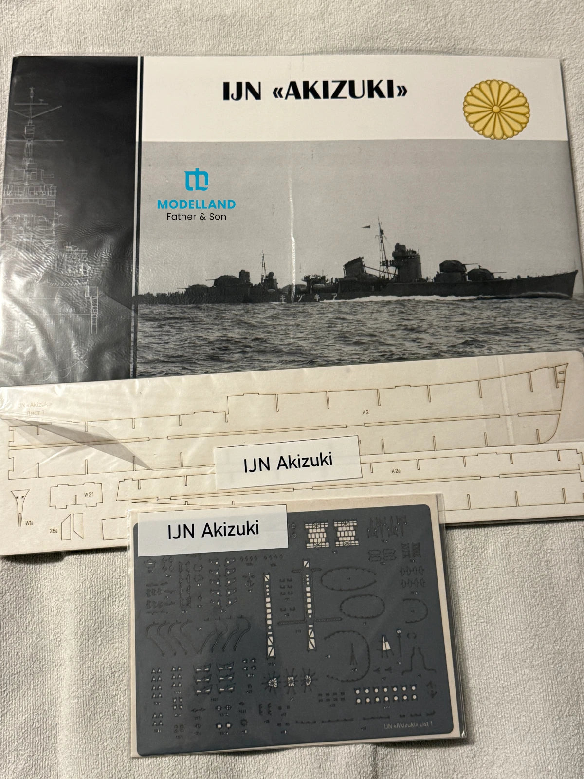 Model kit packaging and accessories laid out on a light fabric surface, including a Modelland Father & Son IJN – Akizuki booklet, a large printed panel, a smaller decal sheet, and a label card.