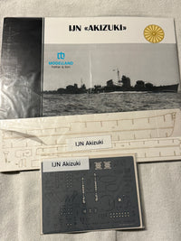 Model kit packaging and accessories laid out on a light fabric surface, including a Modelland Father & Son IJN – Akizuki booklet, a large printed panel, a smaller decal sheet, and a label card.