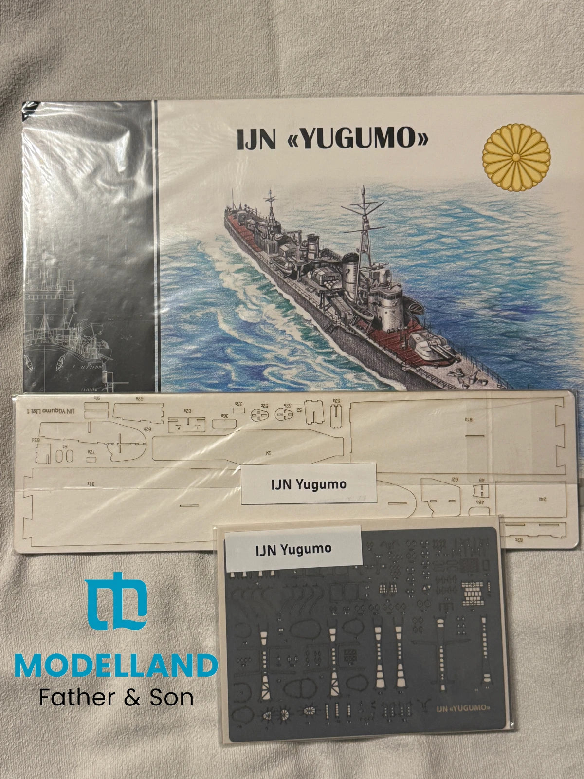 Model kit parts and packaging for IJN Yugumo, including a printed box cover with a navy ship illustration, two flat laser-cut sheet pieces, and a dark gray decal sheet, arranged on a light fabric background. Visible branding includes the blue 'Modelland Father & Son' logo and 'IJN Yugumo' text on the product.