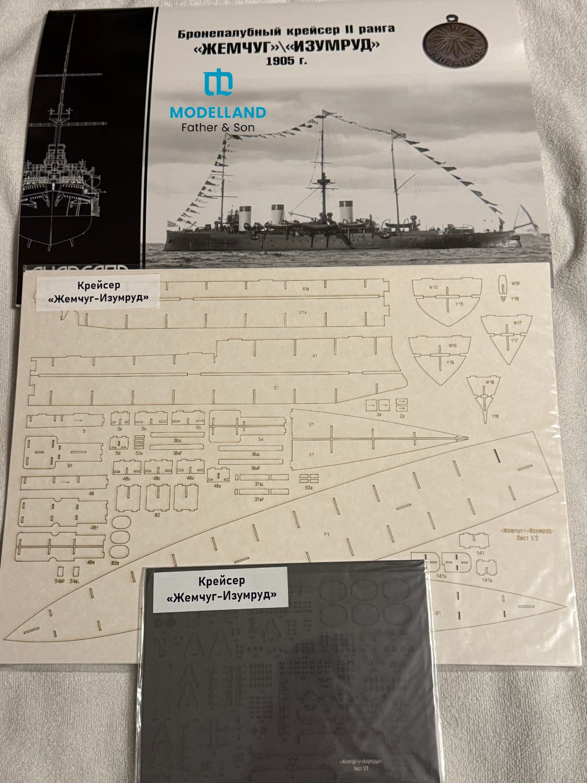 A Modelland model kit package and its contents laid out on a light fabric surface. The set includes a printed cover showing a black-and-white ship illustration, a sheet of decals with technical line drawings, and a small dark gray box labeled in Russian.