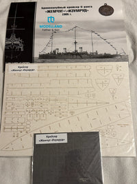 A Modelland model kit package and its contents laid out on a light fabric surface. The set includes a printed cover showing a black-and-white ship illustration, a sheet of decals with technical line drawings, and a small dark gray box labeled in Russian.