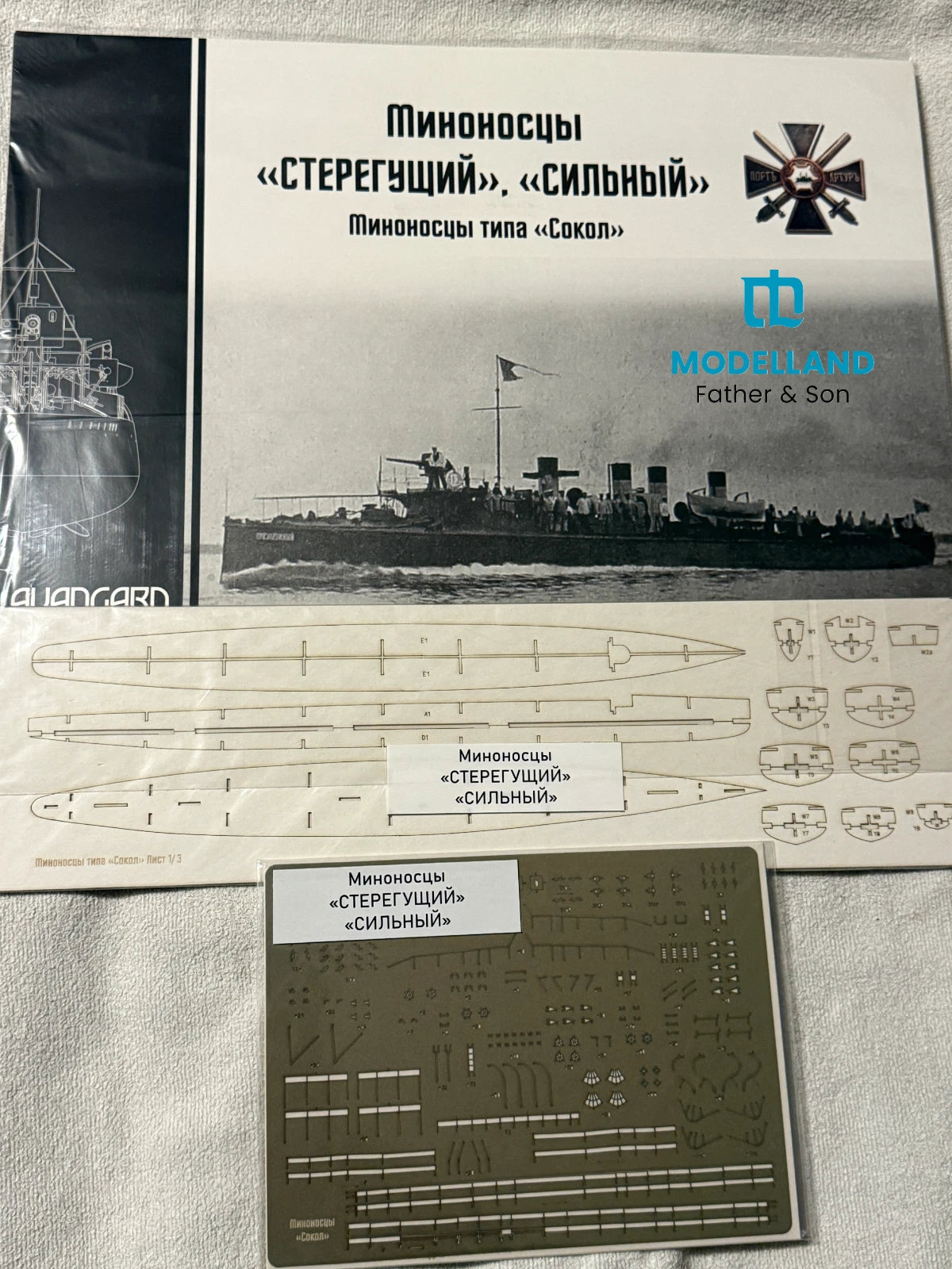 Model kit parts and decals for a Russian submarine, including a printed instruction sheet, a metal decal sheet, and the Modelland Father & Son box cover on a light fabric background.