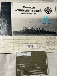 Model kit parts and decals for a Russian submarine, including a printed instruction sheet, a metal decal sheet, and the Modelland Father & Son box cover on a light fabric background.
