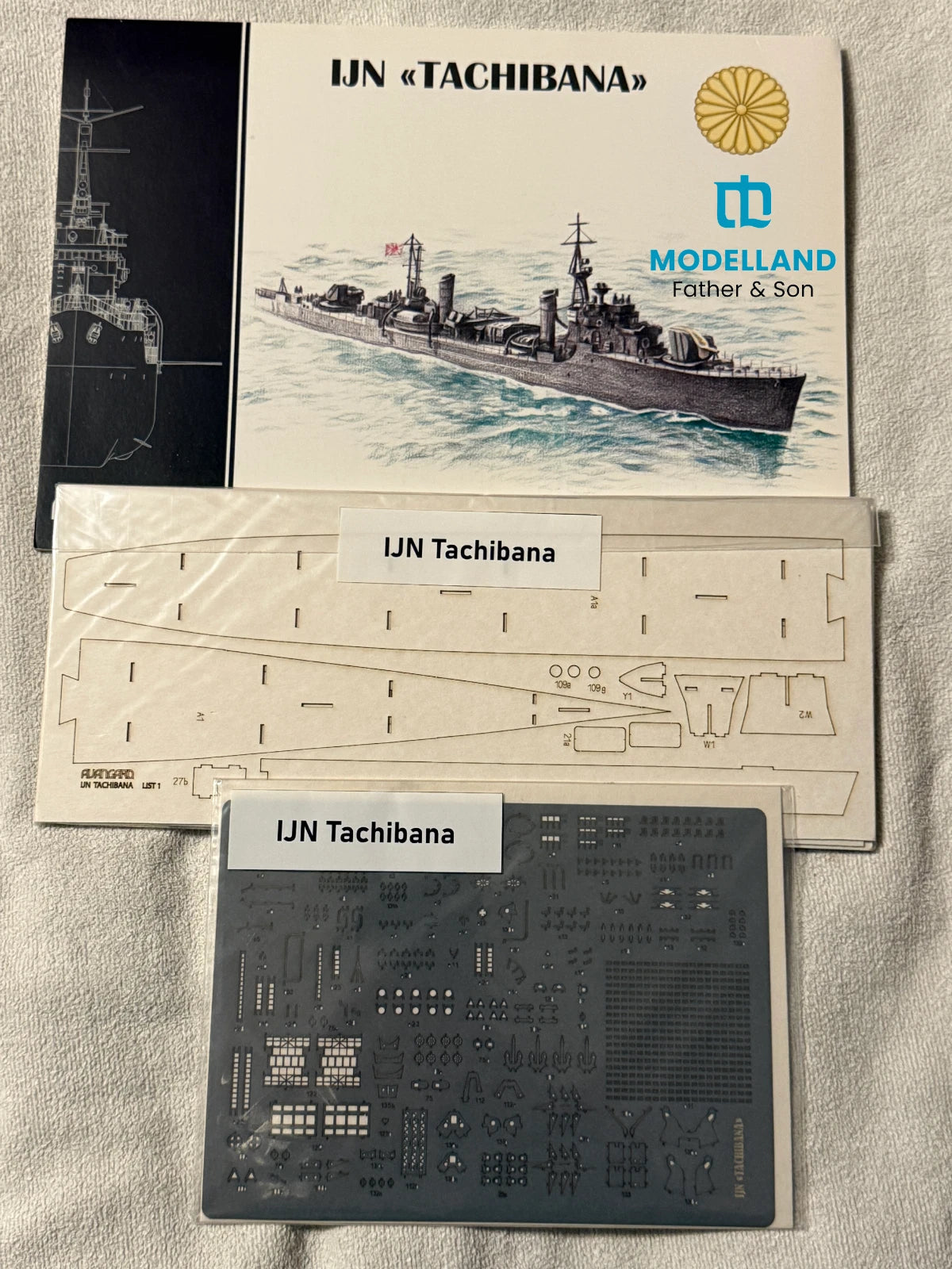 Model kit parts and packaging for IJN Tachibana, including a model ship illustration booklet, a laser-cut sheet, and a gray plastic parts tray on a light fabric background.