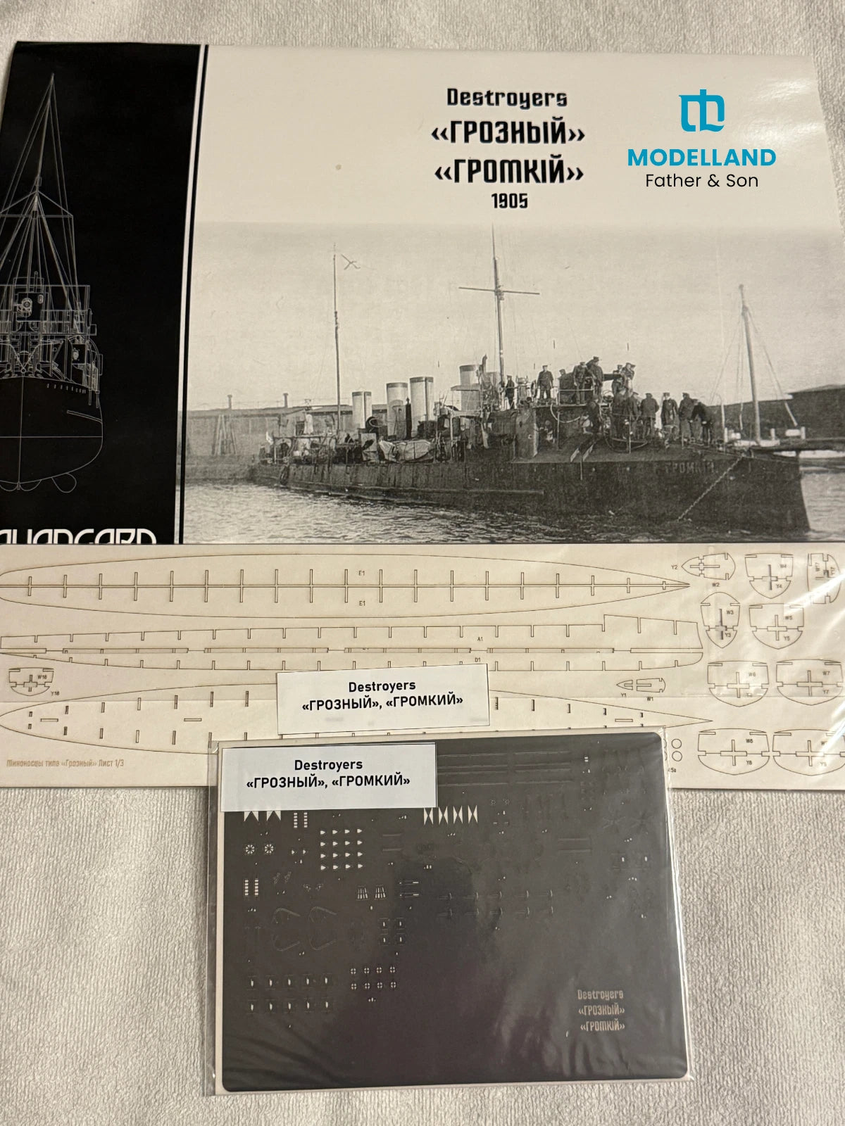 Model kit packaging and accessories for a Modelland Father & Son Destroyers 'Grozny' model ship, including a printed cover with a black-and-white historical photo, scale templates, and a metal sheet.