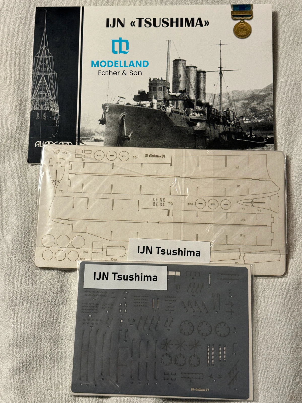 Model kit packaging and accessories laid out on a light fabric background, including a Modelland Father & Son booklet, a printed template sheet, and a gray decal sheet labeled IJN Tsushima.