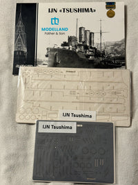 Model kit packaging and accessories laid out on a light fabric background, including a Modelland Father & Son booklet, a printed template sheet, and a gray decal sheet labeled IJN Tsushima.