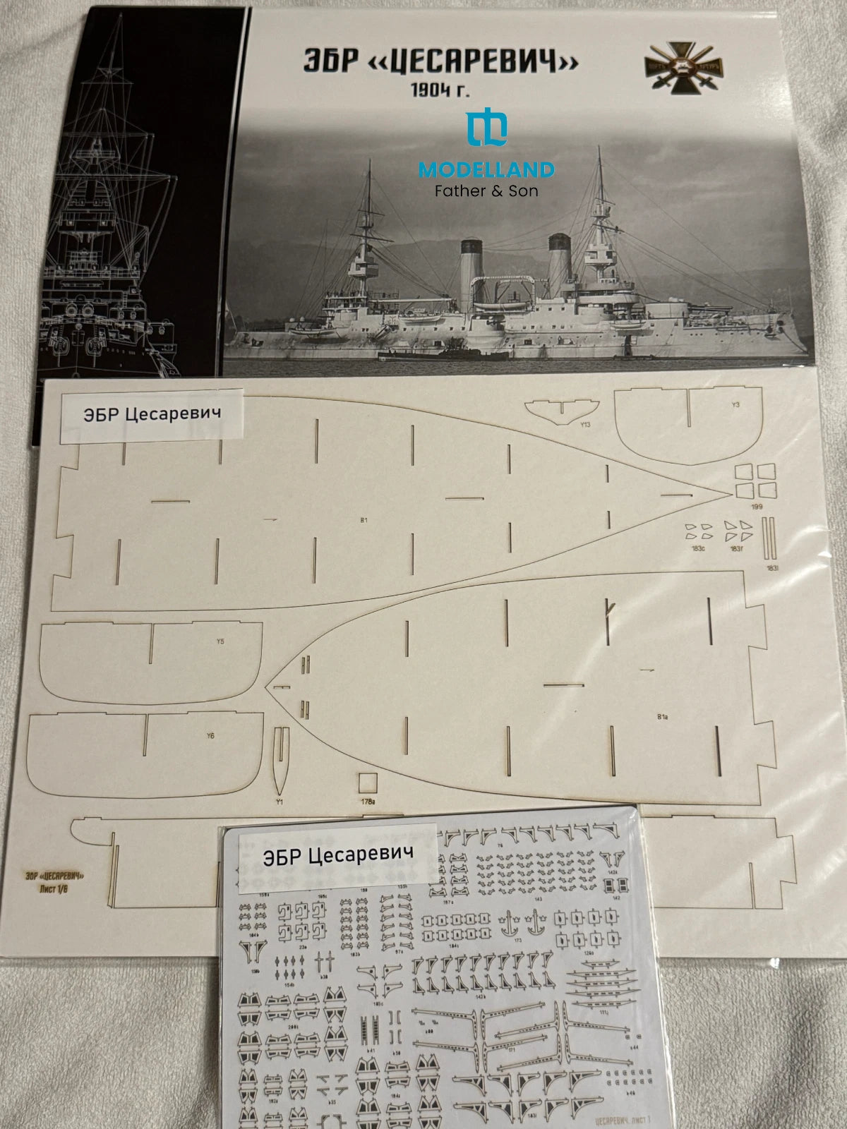 A model kit parts kit for the Russian battleship Tsar Peter I on a light fabric background. The kit includes a printed instruction sheet, a sheet of laser-cut plastic pieces, and a sheet of decals.